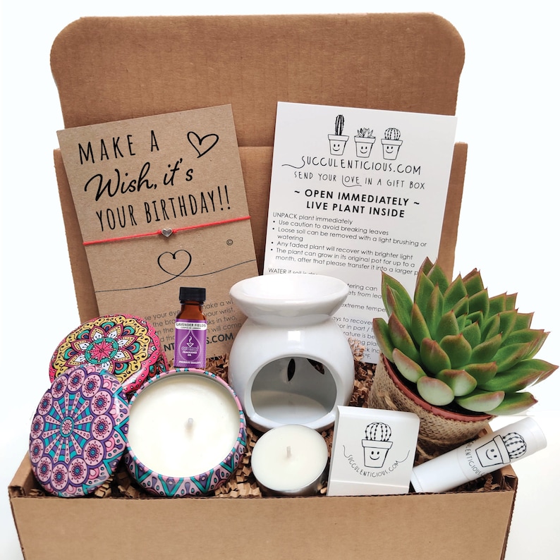 May include: A brown cardboard box filled with a birthday gift set. The set includes a small succulent plant, a candle, a candle holder, a small bottle of essential oil, a lip balm, and a card that says "Make a Wish, it's Your Birthday!"