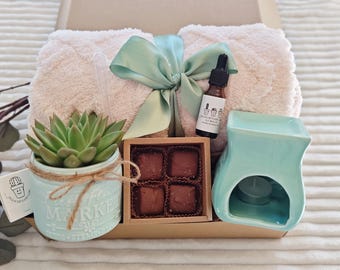 May include: A gift box with a light pink plush blanket, a succulent in a blue pot, chocolates, a teal oil diffuser, and a bottle of essential oil. The box is tied with a light green ribbon.
