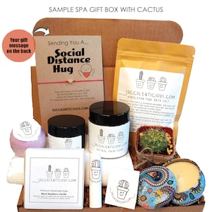 Op de afbeelding: Een bruine kartonnen cadeaudoos gevuld met spa-artikelen, waaronder een badbom, zeep, een suiker scrub, badzout, een kaars en een kleine cactus in een pot. De doos heeft een kaart met de tekst "Sending You A... Social Distance Hug" en een bericht op de achterkant.