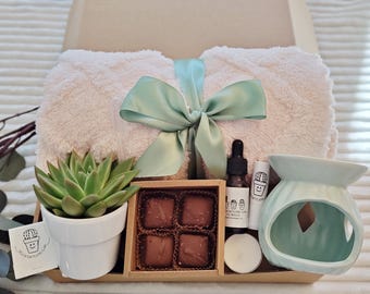 May include: A gift box with a light pink blanket tied with a mint green ribbon. It includes a succulent in a white pot, chocolates, essential oil, lip balm, a candle, and a teal essential oil diffuser.