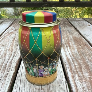 May include: A vintage tin container with a colorful striped lid and a scene of people in a hot air balloon. The container is decorated with a black and white checkered pattern and gold accents.