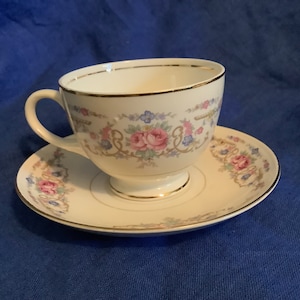 May include: A white teacup and saucer with a floral pattern in shades of pink, blue, and green. The cup and saucer have a gold trim.