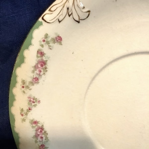 May include: A white porcelain saucer with a green and white scalloped edge. The saucer has a delicate floral pattern in pink and green. The edge of the saucer is decorated with a gold design.