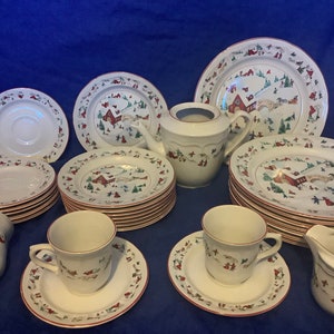May include: A set of white ceramic Christmas dinnerware with a red and green border. The set includes plates, cups, saucers, a teapot, and a creamer. The plates feature a winter scene with a red house, trees, and people.