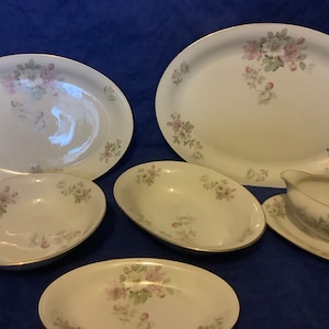 May include: A set of white porcelain serving dishes featuring a floral design in pink, green, and grey. The collection includes oval platters, bowls, and a gravy boat with a gold rim. The items are displayed on a blue surface.