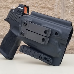May include: A black handgun with a red dot sight in a black holster with a clip on the side.