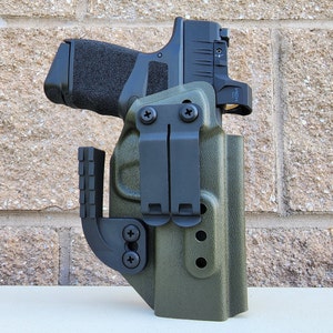 May include: A green and black gun holster with a black clip and a black gun inside. The holster has a textured surface and a black strap.