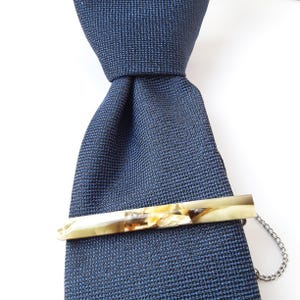 Könnte beinhalten: Eine dunkelblaue Krawatte mit einem goldenen und braunen Krawattenclip. Der Krawattenclip hat eine Kette.