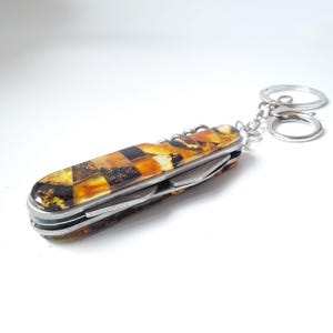 May include: A multi-tool keychain with a decorative handle. The handle features a mosaic pattern of amber, brown, and black tones. The tool includes a corkscrew and other implements. The keychain has two silver rings.