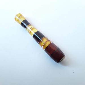 May include: A cylindrical cigarette holder featuring alternating segments of translucent golden amber-colored material and opaque dark brown bands. The smooth, polished surface displays a repeating pattern of light and dark sections along its length.
