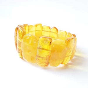 Könnte beinhalten: Ein elastisches Armband bestehend aus mehreren rechteckigen und abgerundeten bernsteinfarbenen Harzsegmenten. Jedes Segment weist einen durchscheinenden, goldgelben Farbton mit sichtbaren inneren Texturen und Variationen auf.