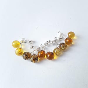 Può includere: Una collezione di orecchini a lobo in ambra con perni in argento. Le sfere di ambra variano dal giallo chiaro al marrone scuro, alcune con inclusioni. Gli orecchini sono esposti su una superficie bianca.