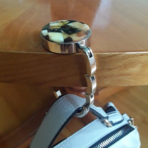 May include: A silver purse hook with a decorative top featuring a mosaic pattern of beige, brown, and black pieces. The hook is attached to a light blue handbag with a black zipper.