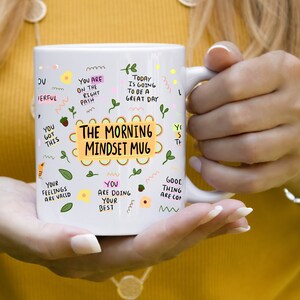 May include: White ceramic mug with a yellow and pink design. The mug has the text "The Morning Mindset Mug" in a yellow box. The mug also has other positive affirmations written on it.