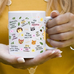 May include: White ceramic mug with a motivational quote design. The mug features illustrations of animals and flowers. The text on the mug includes phrases like "You are capable", "Love gives you strength", "You are powerful", "You got this", "It's okay to make mistakes", and "Your feelings are valid".
