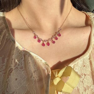 May include: A gold necklace with clear beads and pink, teardrop-shaped gemstones. The necklace is worn over a cream lace top, with a yellow bow. The gemstones are a vibrant shade of pink.