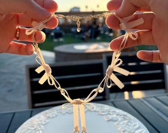 May include: A delicate white ribbon necklace with small bows and crystal accents. The necklace is held in the hands of a person.