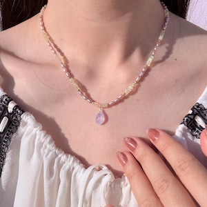 May include: Delicate necklace featuring a strand of small, pink and yellow beads. A small, light purple pendant hangs from the center. The necklace is worn against a white blouse.