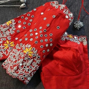 May include: A vibrant red blouse with intricate embroidery and mirror embellishments. The sleeves feature detailed floral and leaf patterns in silver and yellow thread. The blouse has a round neckline and is made of a smooth, shiny fabric.