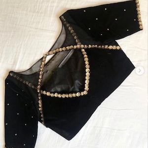 May include: Black velvet top with gold embellishments. The top features a square neckline and sheer mesh detailing. Gold beads are scattered across the sleeves and around the edges. The garment is displayed on a white surface.