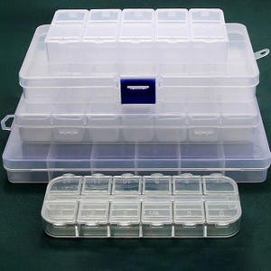 Könnte beinhalten: Transparente Kunststoff-Aufbewahrungsboxen mit mehreren Fächern. Die Behälter sind gestapelt, mit einem kleineren oben und einem kleineren, abgerundeten davor. Der größte Behälter hat einen blauen Verschluss. Ideal zum Organisieren kleiner Gegenstände.