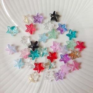 May include: A selection of small, star-shaped beads in a variety of colours, including clear, red, blue, green, purple, and black. The beads are scattered on a white surface, highlighting their translucent and glossy appearance. These decorative beads are suitable for crafting and jewellery making.