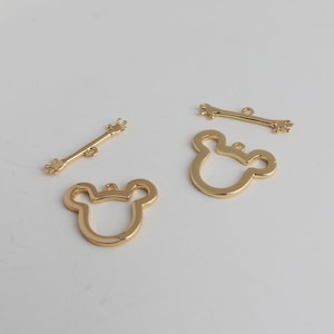 May include: Four gold-colored metal charms, two are shaped like a cartoon mouse head with a hole for hanging, and two are long and thin with a hand shape on each end.