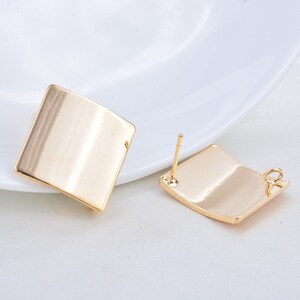 May include: Two gold-colored square stud earrings. The earrings have a smooth, reflective surface and a post for insertion. The earrings are displayed on a white surface.