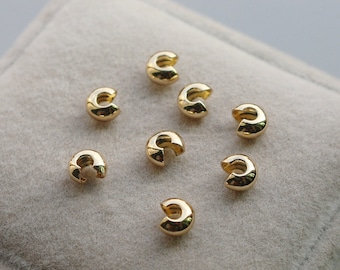 May include: Eight gold-colored metal crimp beads, also known as crimp covers, are displayed on a light beige surface. These beads are used to secure jewelry findings and create a professional finish.
