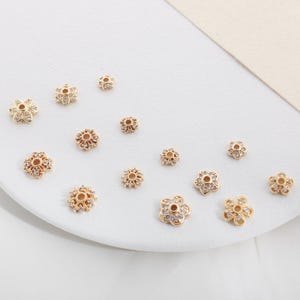 May include: A collection of gold-toned flower-shaped bead spacers with clear rhinestones. The spacers are arranged on a white surface.