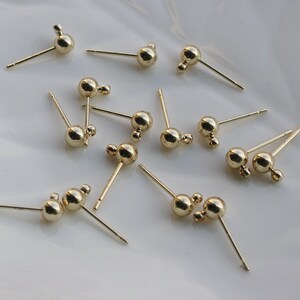 Peut inclure: Un groupe de 10 tiges de boucles d'oreilles dorées avec une petite boule à l'extrémité de chaque tige. Les tiges sont éparpillées sur une surface blanche.