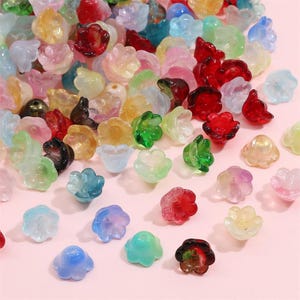 Può includere: Una collezione di perline traslucide a forma di fiore in vari colori, tra cui rosso, verde, blu e giallo. Le perline sono sparse su una superficie rosa, creando un'esposizione colorata e decorativa.