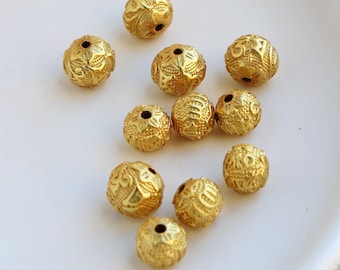 May include: A collection of golden, intricately designed beads. Each bead is spherical, featuring detailed floral and geometric patterns. The beads have a metallic sheen and are scattered on a white surface, ideal for jewelry making or craft projects.
