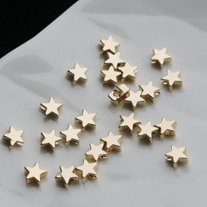 May include: A collection of small, gold-colored star-shaped beads. The beads are scattered on a white surface. Each star has five points and a small hole for stringing. The beads have a shiny, metallic finish.