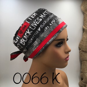 May include: A grey and white scrub hat with a red trim. The hat has the words "Black Lives Matter" printed on it in black and white. The hat is tied with a red ribbon.