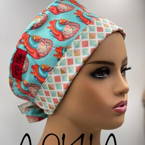 May include: A turquoise scrub hat with a white and pink geometric pattern on the brim. The hat features a repeating pattern of anatomical hearts on a turquoise background. The text "BB Style" is visible on the hat.