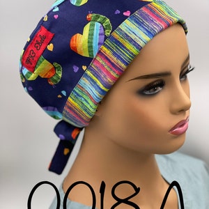 May include: A blue scrub hat with a colorful rainbow heart and caterpillar print. The hat has a rainbow striped trim and a tie at the back. The text "Miss Eble" is visible on the hat. The hat is labeled "0018 A".