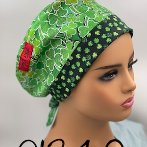 Puede incluir:  gorro de quir&oacute;fano verde con estampado de tr&eacute;bol. El gorro tiene un ribete negro y verde y una corbata en la parte posterior. El gorro est&aacute; etiquetado como "Belle Scrubs".