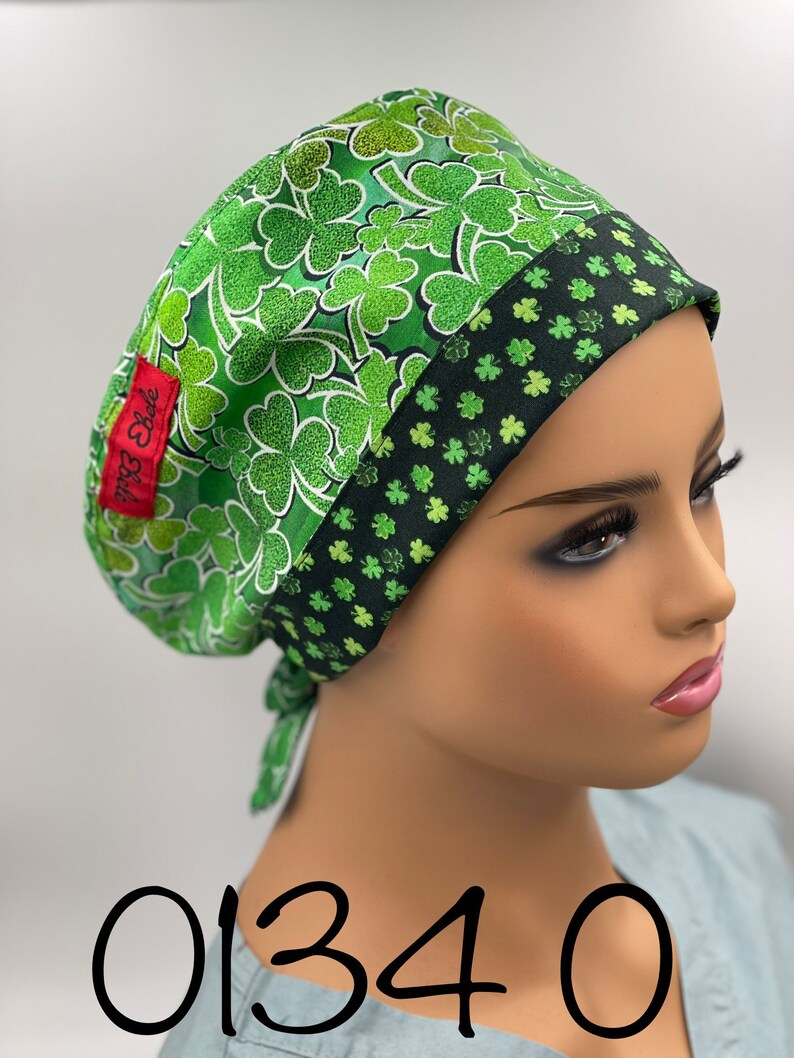 Puede incluir:  gorro de quir&oacute;fano verde con estampado de tr&eacute;bol. El gorro tiene un ribete negro y verde y una corbata en la parte posterior. El gorro est&aacute; etiquetado como "Belle Scrubs".
