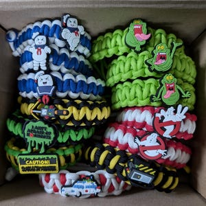 Op de afbeelding: Een verzameling kleurrijke paracord armbanden met verschillende Ghostbusters-themacharms, waaronder Slimer, de Stay Puft Marshmallow Man en het Ghostbusters-logo. De armbanden zijn gemaakt van groene, rode, witte, zwarte en gele paracord.