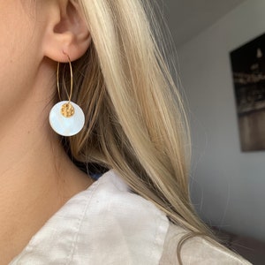 May include: Gold hoop earrings with a white shell pendant. The shell has a textured surface and a gold accent.