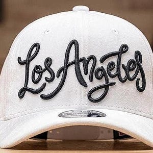 May include: A white baseball cap featuring the embroidered text "Los Angeles" in black cursive lettering. The cap has a curved peak and a button at the crown. The background is blurred, implying a workshop environment.