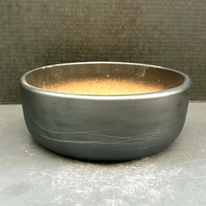 Puede incluir: Un cuenco pequeño y redondo de cerámica negra con un exterior liso y un interior marrón claro contrastante. El cuenco está sobre una superficie gris oscuro.