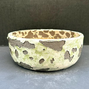 May include: A small, round ceramic bowl with a distressed finish. The bowl is painted in a pale green color with dark brown accents. The rim and exterior have a textured, chipped appearance.