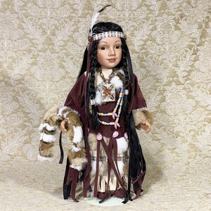 May include: A porcelain doll dressed in a brown suede dress with a faux fur trim. The doll has long black hair, brown eyes, and is wearing a beaded necklace and a headband with feathers.