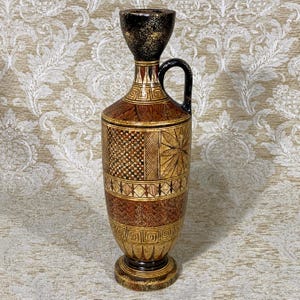 May include: A decorative, hand-painted vase with a black handle and a flared top. The vase features intricate geometric patterns in shades of brown, gold, and black. The base is round and the vase is approximately 14 inches tall.