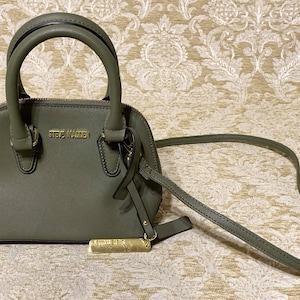 May include: A small, olive green satchel handbag with a top handle and a long shoulder strap. The bag has a gold zipper and a gold metal tag with the brand name "Steve Madden" on it.