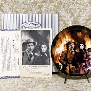 May include: A decorative ceramic plate with a gold rim features a painting of a couple in a burning city. The plate is on a black stand. The text "The Burning of Atlanta" is printed on a white card with a black and white photo of the couple.