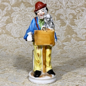 May include: A porcelain figurine of a clown playing a hand-crank organ. The clown is wearing a red hat, blue jacket, and yellow pants. A small monkey sits on the organ.