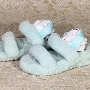 May include: A pair of light blue UGG slides with a fluffy, textured appearance. Each slide features two thick straps and a branded band. The slides are presented on a patterned surface.